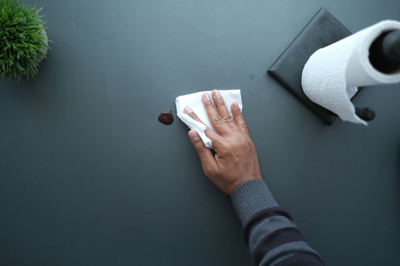 services-02 A hand wipes a dark surface with a tissue near a paper roll and plant.